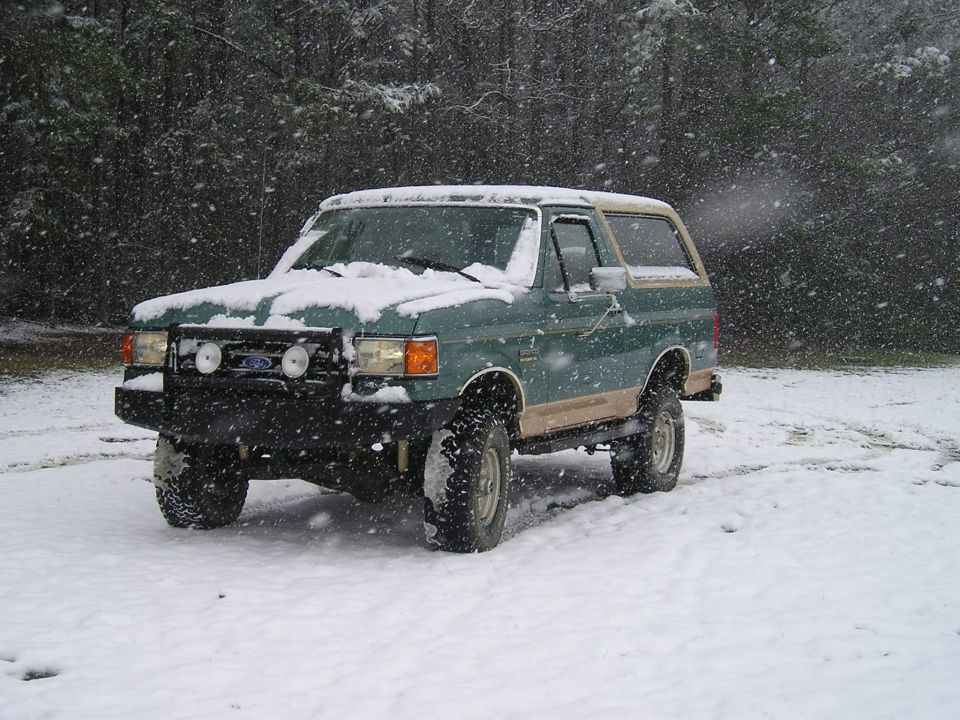 Green and tan Ford Bronco parked in snow with falling snow.