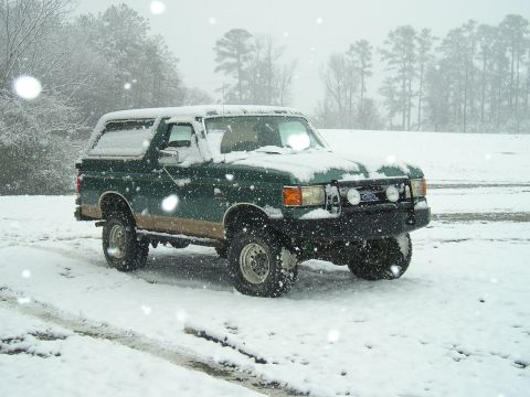 1989 Ford Bronco - A dark green Ford Bronco with a tan stripe sits in a snowy field during a snowfall. The vehicle has aftermarket lights mounted on the front…