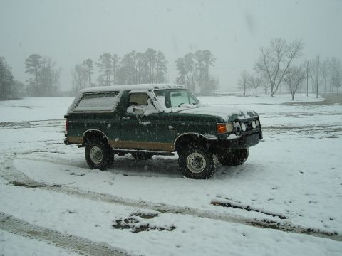1989 Ford Bronco - A dark green Ford Bronco is parked in a snowy field during a snowfall. The vehicle has a two-tone paint scheme with a brown lower body pane…