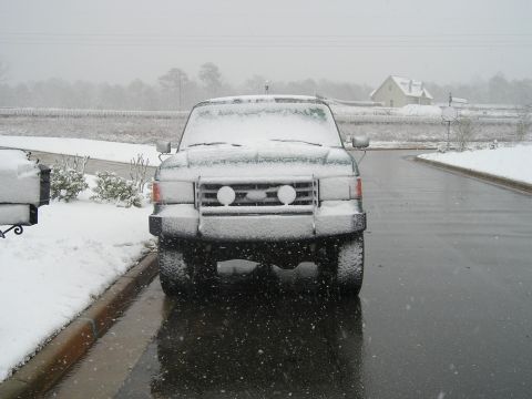 1989 Ford Bronco - A snow-covered Ford Bronco is parked on a wet asphalt road during a snowfall. The front of the vehicle is visible, with snow accumulating o…