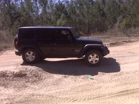 2008 Jeep Wrangler - A black four-door Jeep Wrangler is parked on a dirt road with a line of trees in the background. The vehicle is viewed from the side, showi…