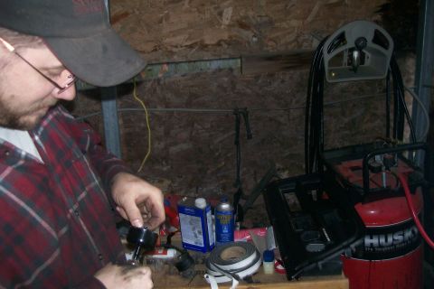1982 Ford E350 - A person wearing a red plaid shirt and a baseball cap is working on a small black object. Various tools and supplies are visible on a woode…