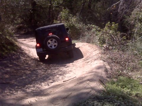 2008 Jeep Wrangler - A black Jeep Wrangler is shown from the rear driving on a dirt road through a wooded area. The vehicle is tilted to the right as it navigat…