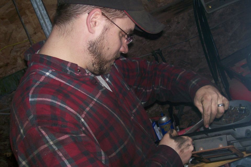 Person in a red plaid shirt working with dirty hands, wearing glasses and a cap.