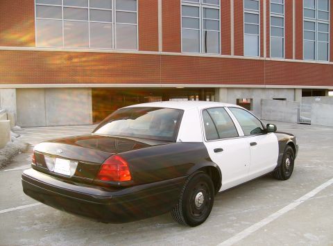 2004 Ford Crown Victoria - A two-tone police car, with a black rear and white front, is parked in a lot with a brick building in the background.