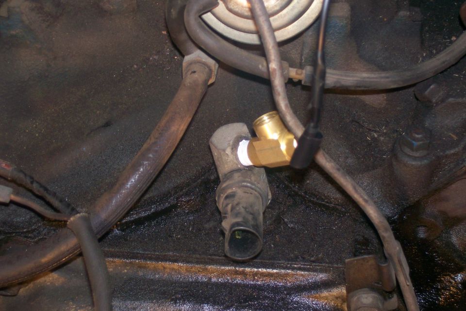 Close-up of an engine block showing a brass sensor adapter installed on a threaded port, with hoses and wiring nearby