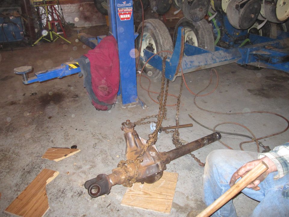 Rusty differential axle assembly suspended by chains in a garage, with a person holding a wooden handle nearby