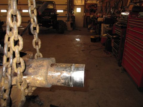 1992 Ford Bronco - A rusty axle part hangs from chains in a workshop, with a modified off-road vehicle visible in the background.