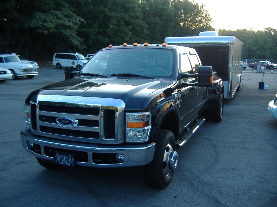 Black Ford Super Duty truck with dual rear wheels towing a silver enclosed trailer. Other vehicles visible in background