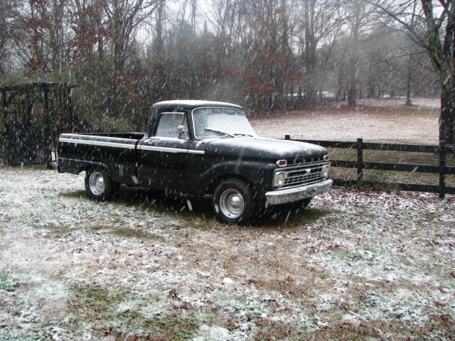 Black pickup truck parked in a snowy field with trees and a fence in the background