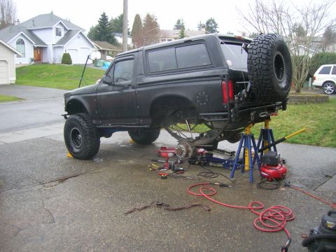 1996 Ford Bronco - Flat-black lifted Bronco raised on blue jack stands in a driveway with the rear axle and both rear wheels removed, exposing the brake backi…