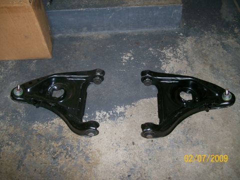 1995 Mercury Grand Marquis - Two black control arms are shown on a concrete floor, possibly for a vehicle's suspension system.