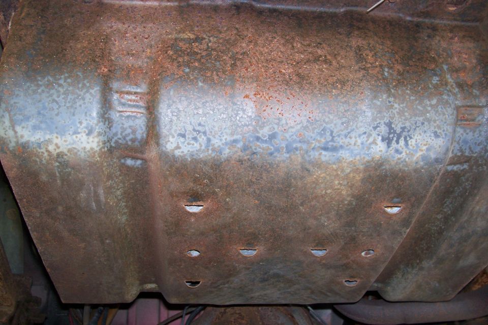 Heavily rusted metal undercarriage part with visible indentations and holes. Some blueish discoloration present