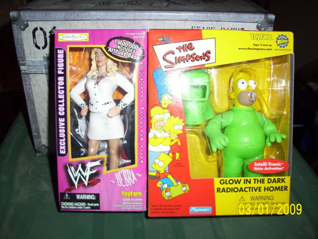 Two toy figures in packaging, one WWF wrestler Debra, the other Radioactive Homer from The Simpsons, on a table