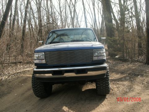 1996 Ford Bronco - Front view of a lifted blue Ford truck with a chrome grille and large tires in a wooded area.