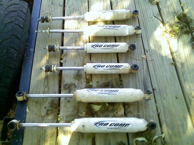 Six Pro Comp shock absorbers laid out on a wooden surface next to a tire