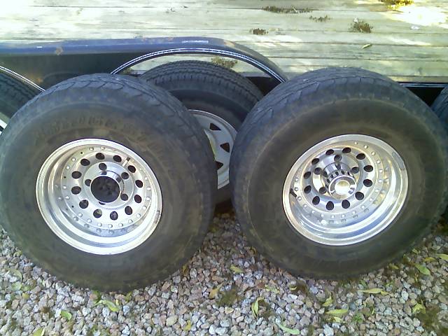 Two aftermarket wheels with tires, resting on gravel next to a trailer