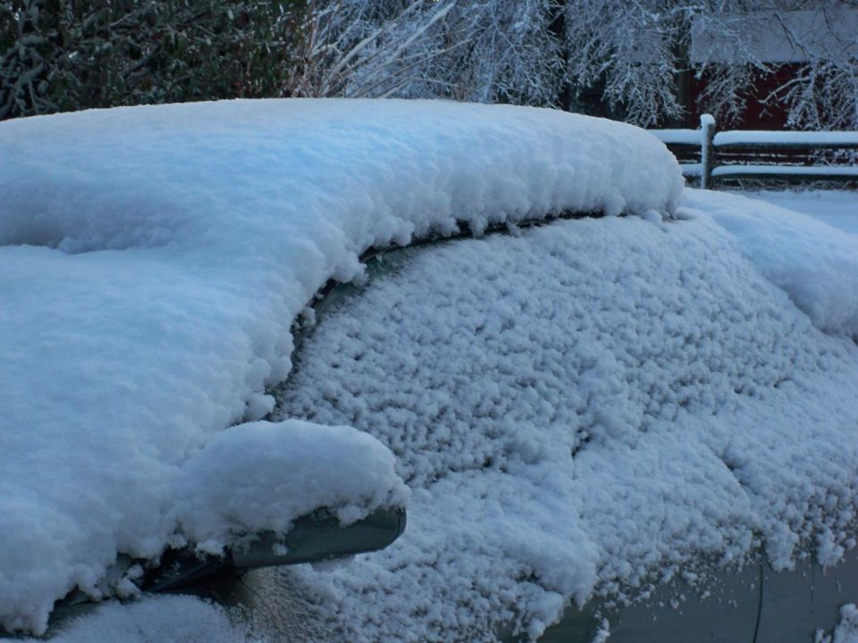 Car roof and side mirror covered in thick snow, with a red barn and fence in the background