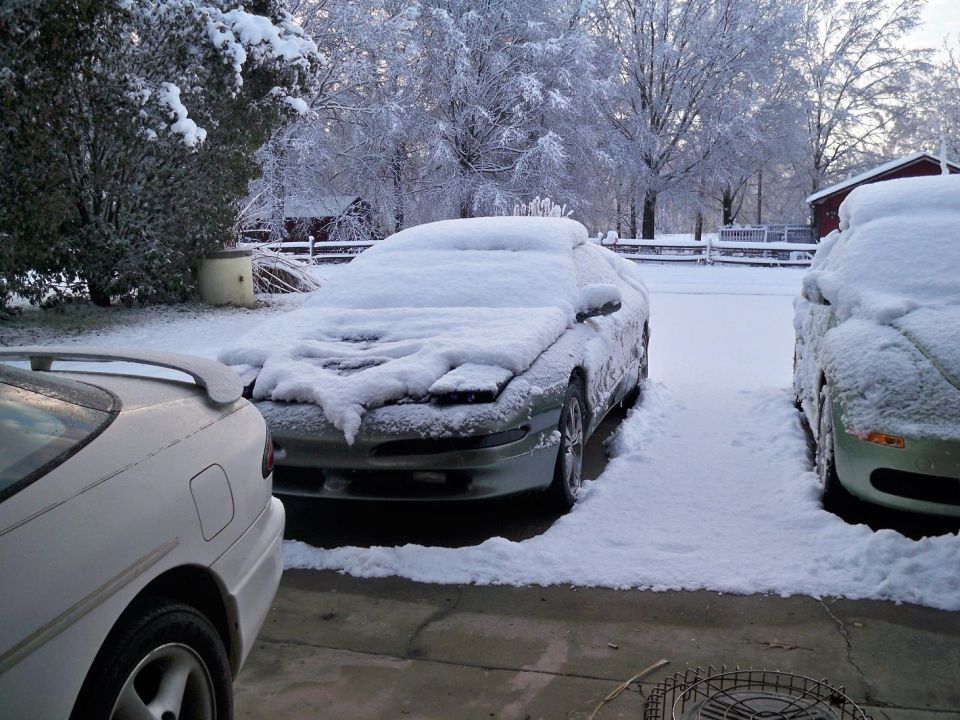 two cars parked outside covered in snow, with trees and a house in the background