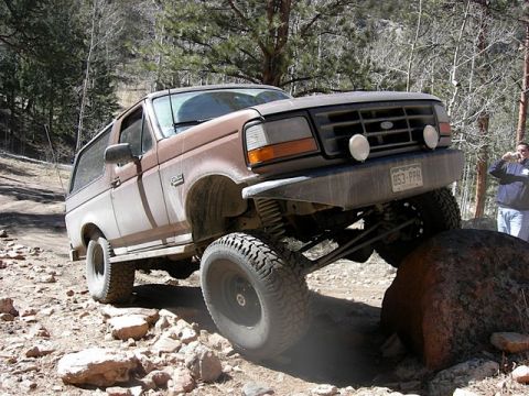 1993 Ford Bronco - This modified Ford Bronco is shown off-roading on a rough, rocky terrain with trees in the background.