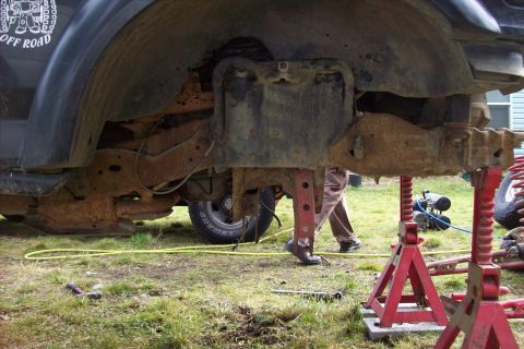 1995 Ford Bronco - The underside of a heavily rusted Ford Bronco chassis is supported by red jack stands in a grassy yard.