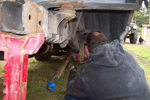 1995 Ford Bronco - Man welding on vehicle chassis while it's on jack stands; another vehicle and dogs are in the background.