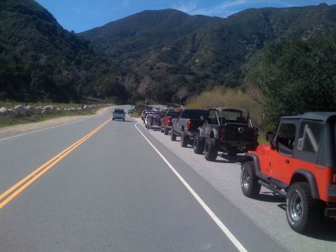 1979 Ford Bronco - A line of off-road vehicles, including a red Jeep Wrangler, are parked on the shoulder of a two-lane road in a mountainous, wooded area.