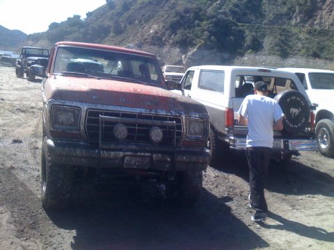 1979 Ford Bronco - A muddy red Ford pickup truck is parked in the foreground, facing left. Behind it, a white Ford Bronco with a spare tire on the back is vis…