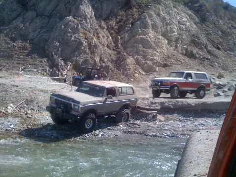 1979 Ford Bronco - Two off-road vehicles are parked on a rocky shore next to a body of water. A brown Ford Bronco is in the foreground, and a red and white Fo…