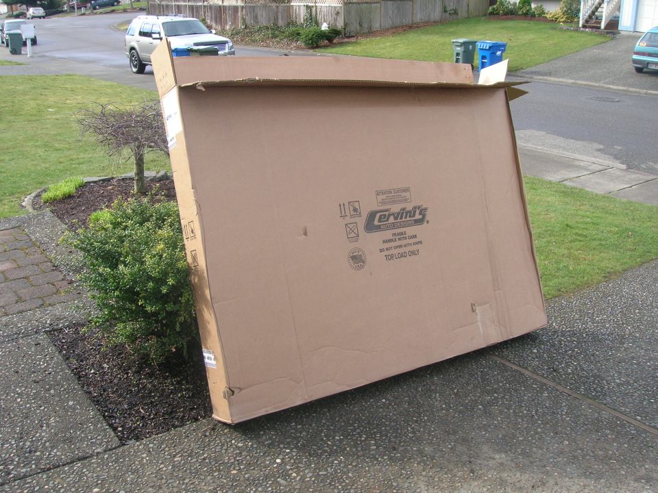 Large cardboard box with Cervini's Auto Designs logo, leaning on a bush, with a silver SUV in the background.