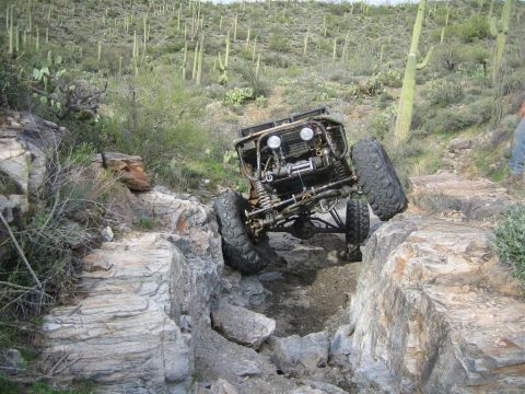 1987 Suzuki Samurai - A heavily modified off-road vehicle navigates a rocky, desert terrain with large saguaro cacti in the background. The vehicle is tilted at…