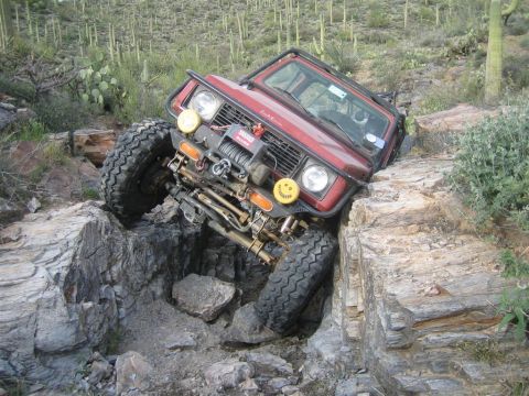 1987 Suzuki Samurai - A red Suzuki Samurai is navigating a steep, rocky off-road trail, with its front tire precariously perched on a large rock.