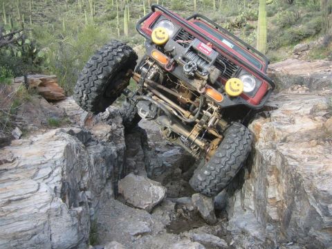 1987 Suzuki Samurai - A red Suzuki Samurai is navigating a steep, rocky incline, with its front tire precariously perched on a rock ledge. The vehicle is heavily…