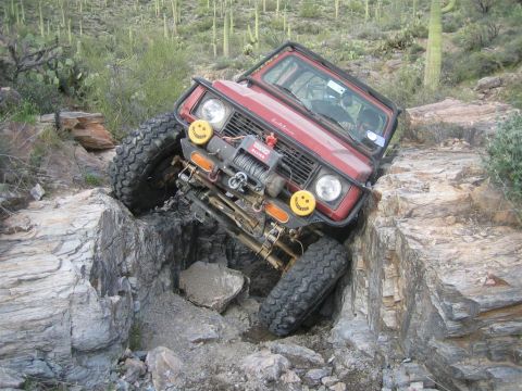 1987 Suzuki Samurai - A red Suzuki Samurai is tilted at a severe angle, with its front right tire wedged in a rocky crevice. The off-road vehicle is navigating a…