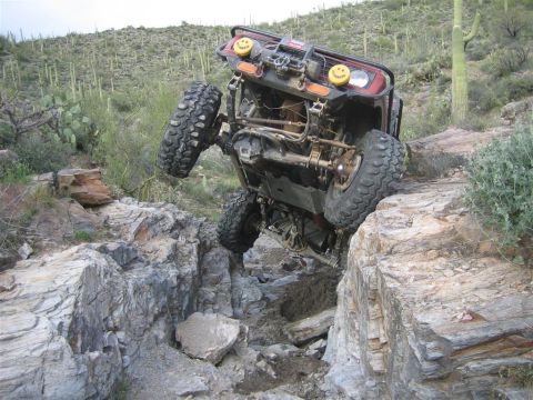 1987 Suzuki Samurai - An off-road vehicle is tilted precariously on its side, with its tires high in the air, navigating a rocky, desert terrain with saguaro cac…