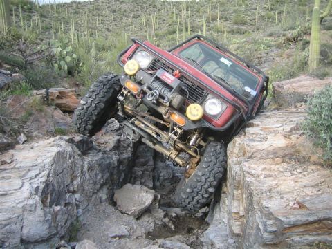 1987 Suzuki Samurai - A red 1987 Suzuki Samurai is navigating a steep, rocky off-road trail, with its front left tire high in the air and its front right tire pr…