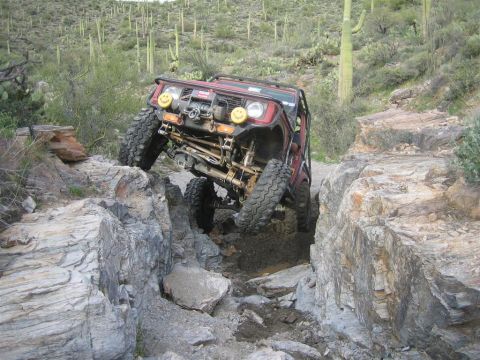 1987 Suzuki Samurai - A red off-road Suzuki Samurai navigates a rocky, desert trail, with its front tire climbing over a large boulder. The vehicle is equipped w…
