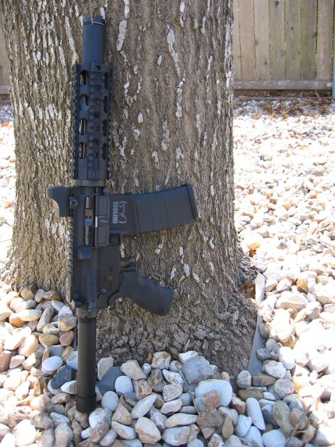 1996 Ford Bronco - A black rifle is leaning against a tree trunk, surrounded by rocks and gravel. The rifle has "INFIDEL" written on the magazine in Arabic sc…