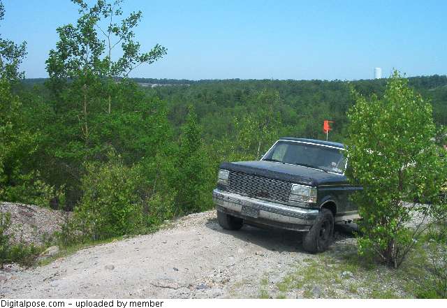 Dark green Ford Bronco with whip flag parked on a gravel trail above woods
