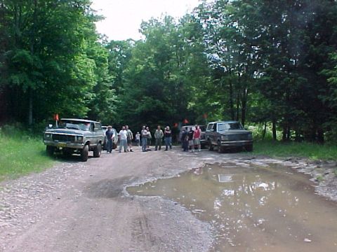 1993 Ford Bronco - A group of people stands between parked 4x4s on a muddy gravel trail cut through dense green woods, with a two-tone lifted pickup on the le…
