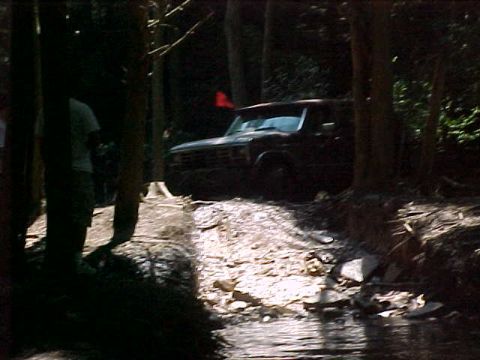 1993 Ford Bronco - A lifted full-size SUV with a red flexible whip flag sits in deep shade on a rocky trail approach beside a creek, viewed at a 3/4-front ang…