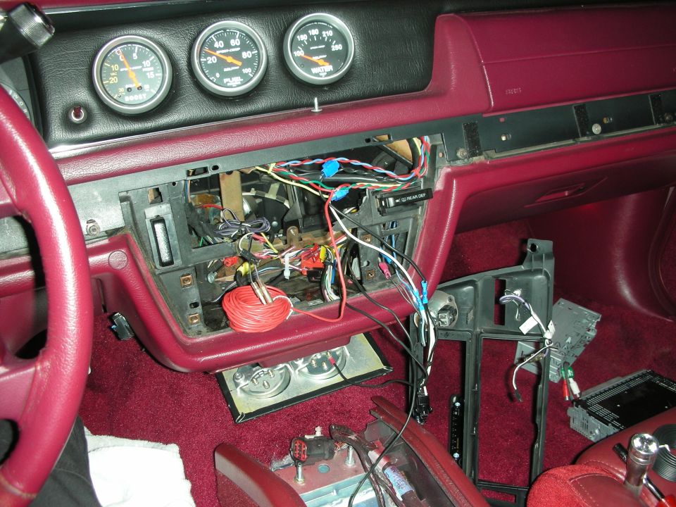 Car interior with disassembled dashboard, wiring, gauges, red steering wheel and carpet.