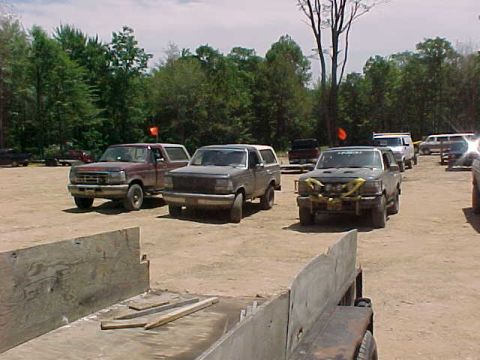 1993 Ford Bronco - A row of mud-covered trucks parked in a dirt staging lot at a wooded off-road park, including a maroon Ford pickup, a full-size Bronco with…