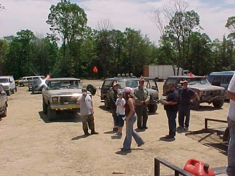 1993 Ford Bronco - Group of people standing and talking in a dirt staging lot at a wooded off-road park, surrounded by parked 4x4 trucks and SUVs with orange…