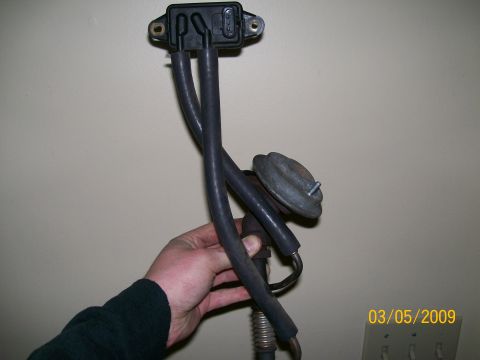 1995 Mercury Grand Marquis - A hand holds a vehicle part consisting of a black sensor unit with three rubber hoses attached. A metal diaphragm actuator is connected to…