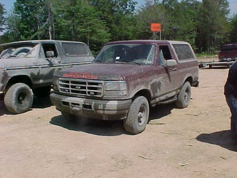 1993 Ford Bronco - A dark red full-size Ford Bronco caked in dried mud sits on a sandy staging area, front three-quarter view with an orange safety flag on a…