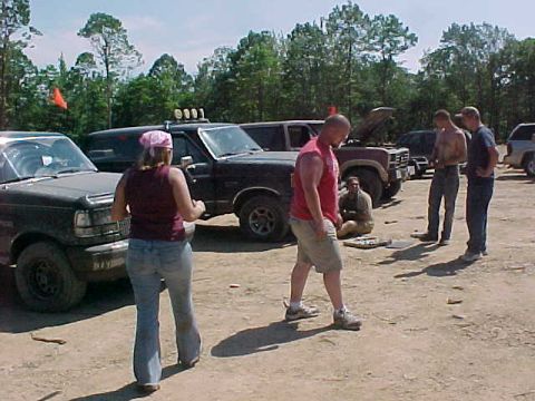 1993 Ford Bronco - A group of mud-covered lifted Ford trucks and Broncos parked in a dirt staging lot at the edge of pine woods, one with its hood up and anot…