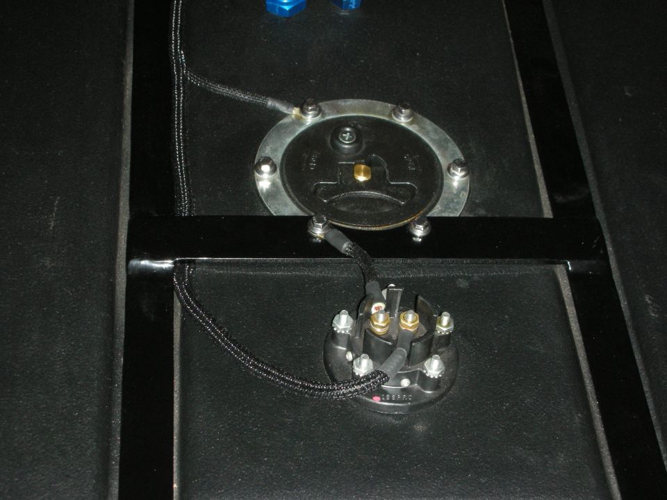 Two black fuel pump modules with braided hoses and wires, connected to a dark surface with a black metal frame.