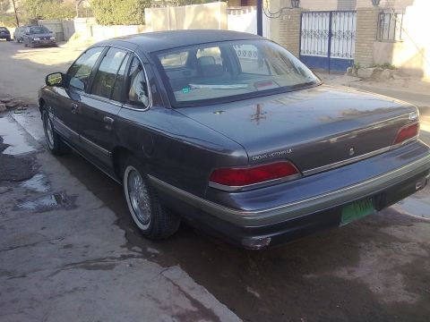 1992 Ford Crown Victoria - Three-quarter rear view of a dark gray two-tone Ford Crown Victoria LX sedan parked on a wet street in front of a walled house with barred…
