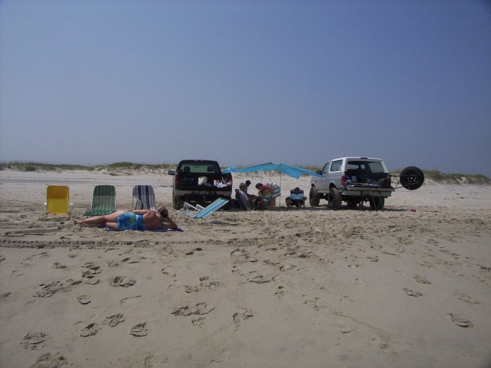 White Ford Bronco and dark pickup parked on beach with tarp canopy, people, and beach chairs in sand